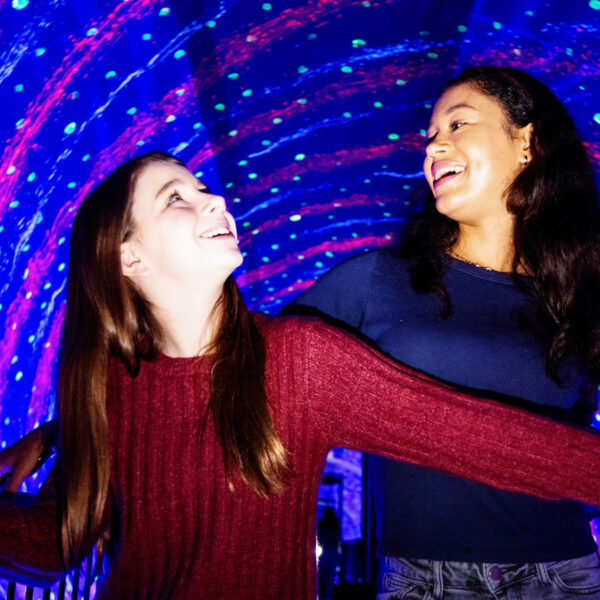 Two friends laugh and hold onto railings as they walk through a vibrant, swirling tunnel of neon lights and optical illusions at the Museum of Illusions in Philadelphia.