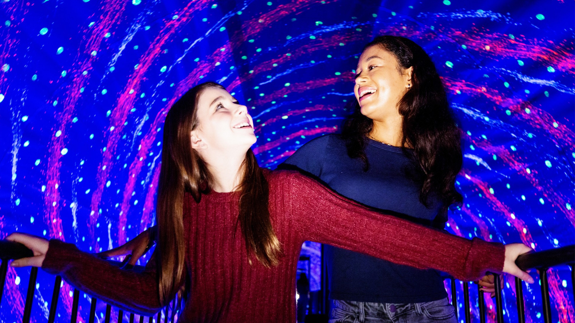 Two friends laugh and hold onto railings as they walk through a vibrant, swirling tunnel of neon lights and optical illusions at the Museum of Illusions in Philadelphia.