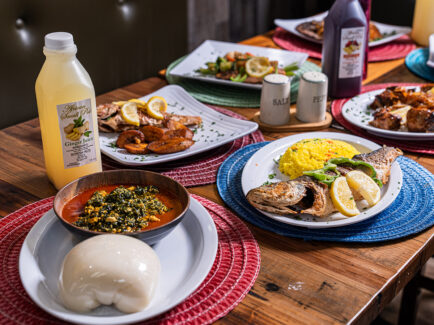 Table spread of traditional West African dishes, including grilled whole fish with rice, egusi soup with pounded yam and fried plantains, alongside bottles of ginger and hibiscus juice.
