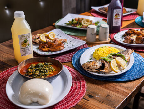 Table spread of traditional West African dishes, including grilled whole fish with rice, egusi soup with pounded yam and fried plantains, alongside bottles of ginger and hibiscus juice.