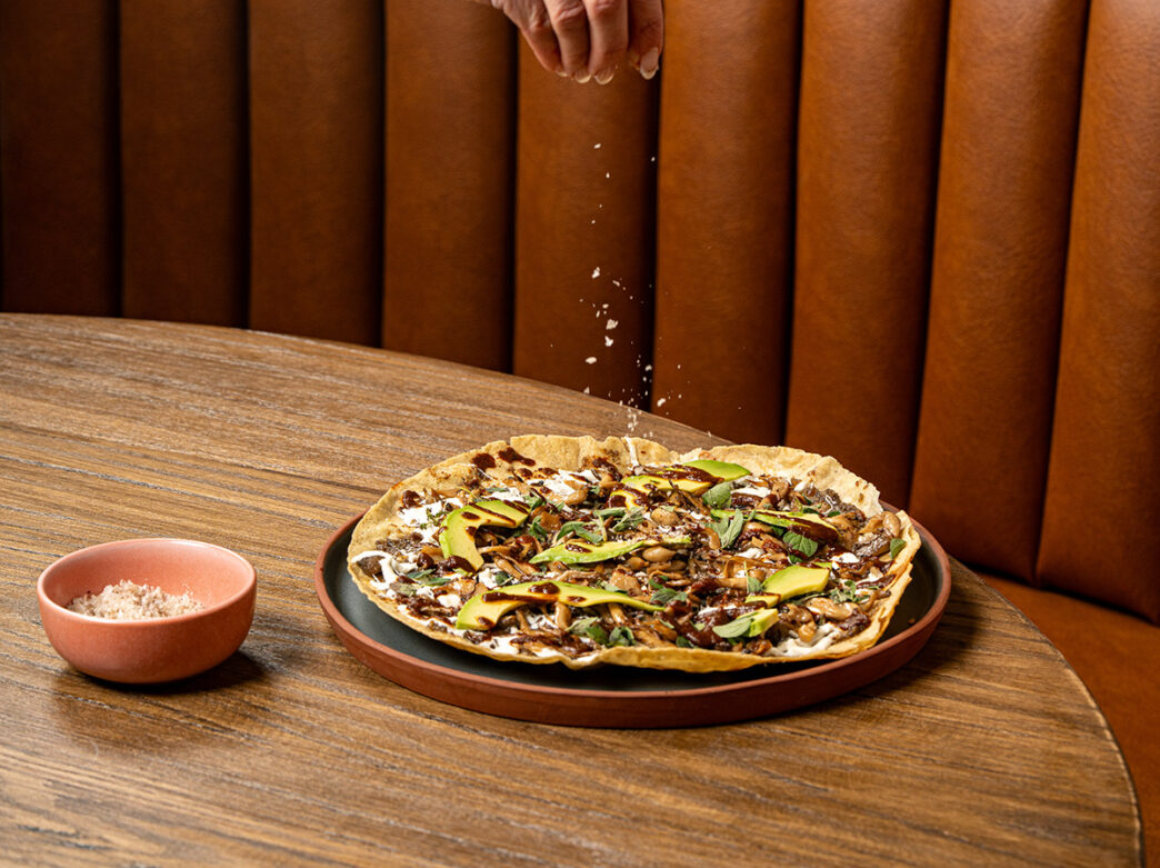 Una mano espolvorea sal sobre una tlayuda de setas que descansa sobre una hermosa mesa de madera.