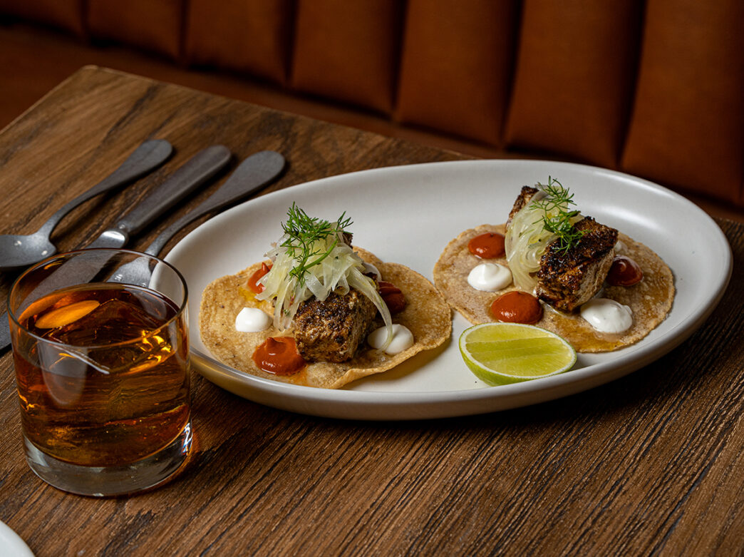 Two swordfish tacos served on a white oval plate with a lime wedge and a cocktail on a wooden table.