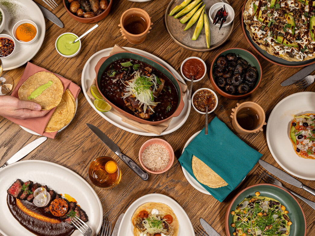 A variety of Mexican dishes, including tortillas, a tuna tostada and carne asada, sit on a wooden table at Amá