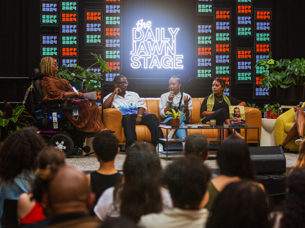 A group of panelists speak on stage at BlackStar Film Festival's Daily Jawn Stage in front of a live audience, framed by plants and vibrant neon sign.