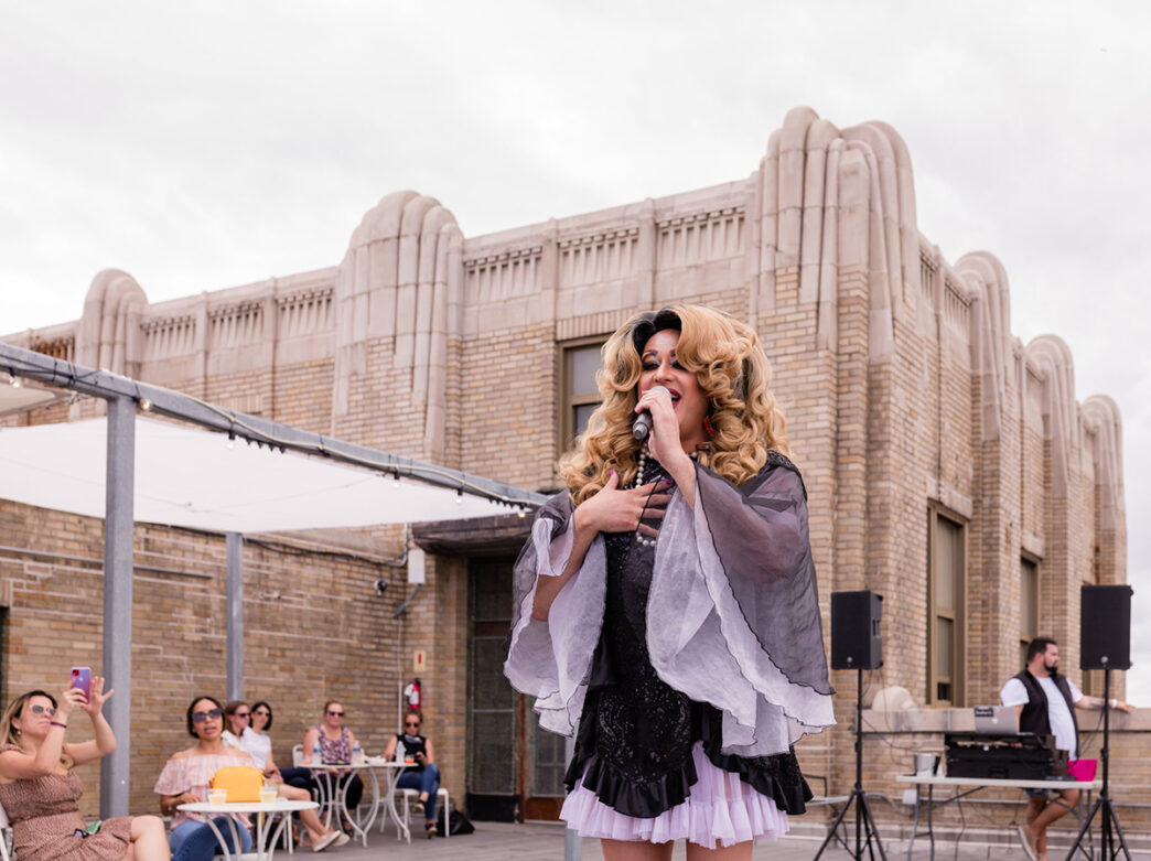 Une travestisseuse glamour chante dans un microphone au Bok Bar, un bar situé sur le toit de la ville. Elle porte une perruque volumineuse et un collier de perles, tandis que les clients assis regardent et prennent des photos.