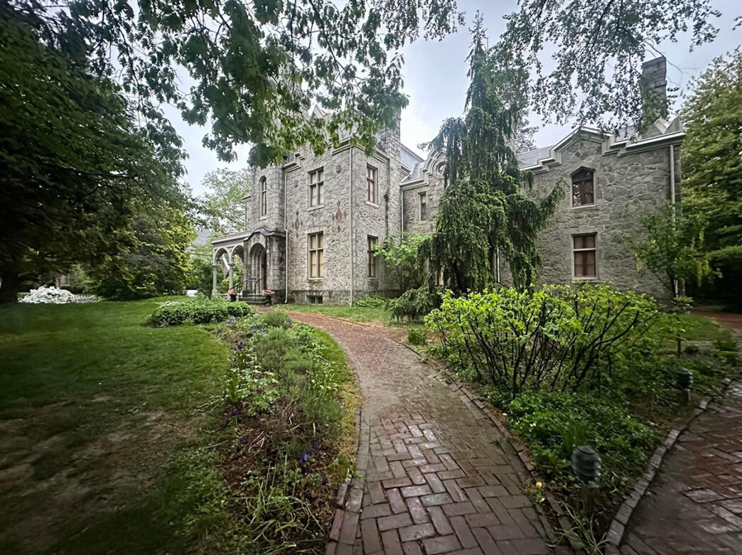A historic gray stone mansion with Gothic-style architecture surrounded by lush greenery, a brick pathway and garden beds.