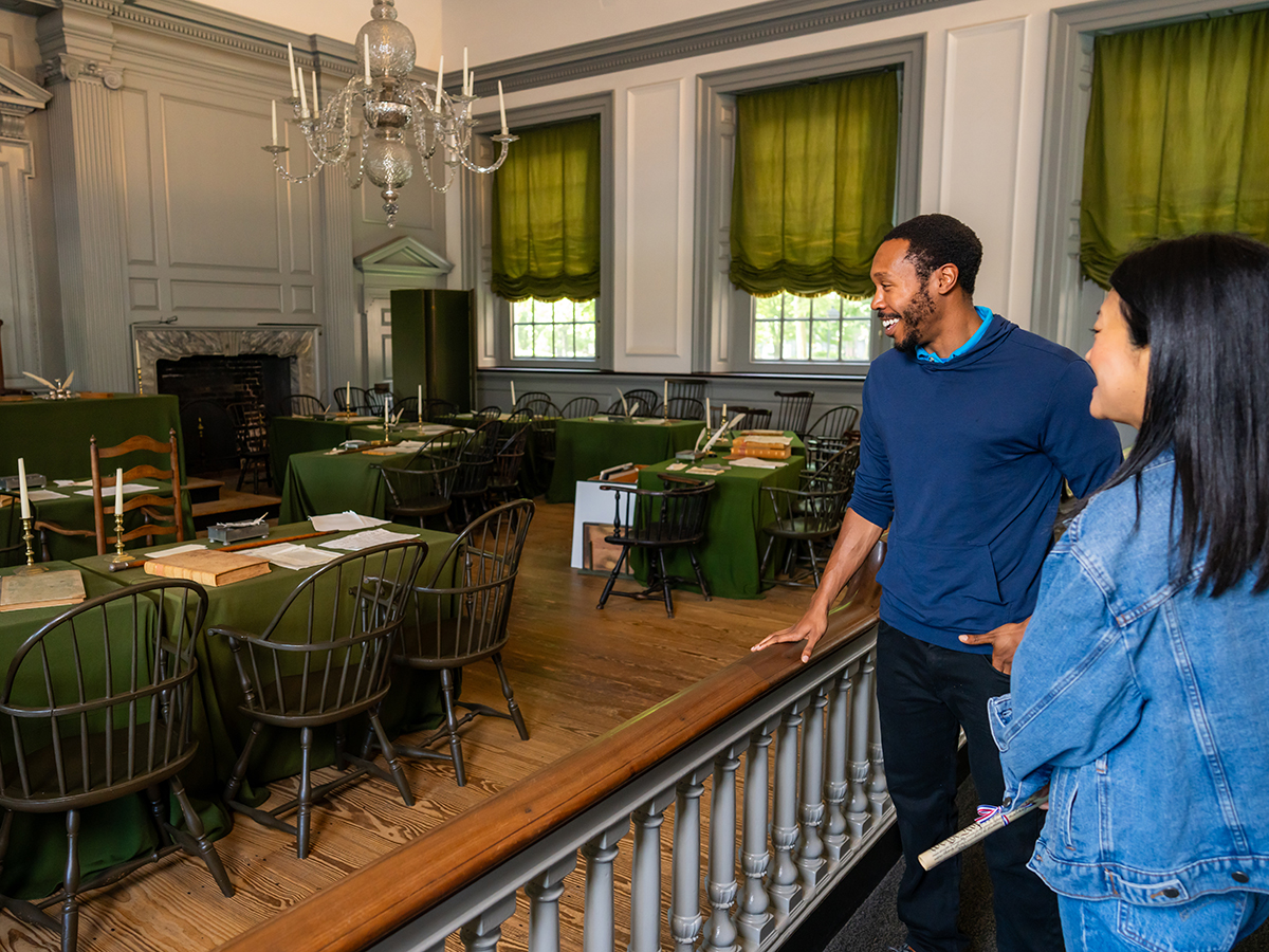 Two visitors tour the interior of the Assembly Room that has green-draped desks and a prominent chandelier above the meeting space.