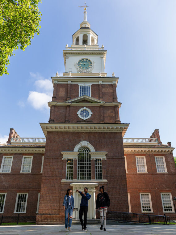 Three people walk with the back of Philadelphia's Independence Hall in the background.