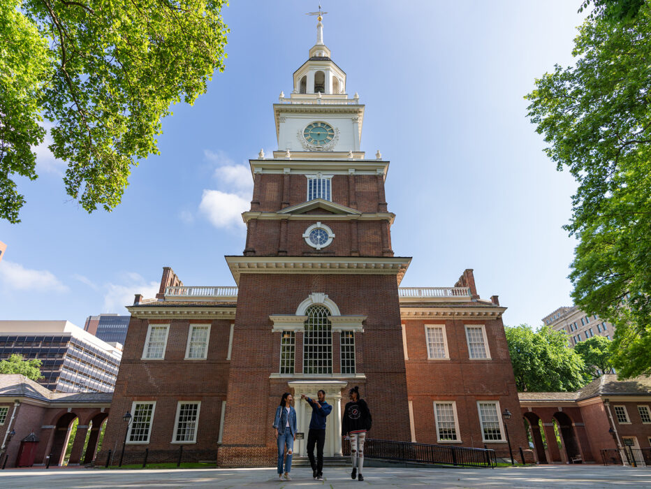 Three people walk with the back of Philadelphia's Independence Hall in the background.
