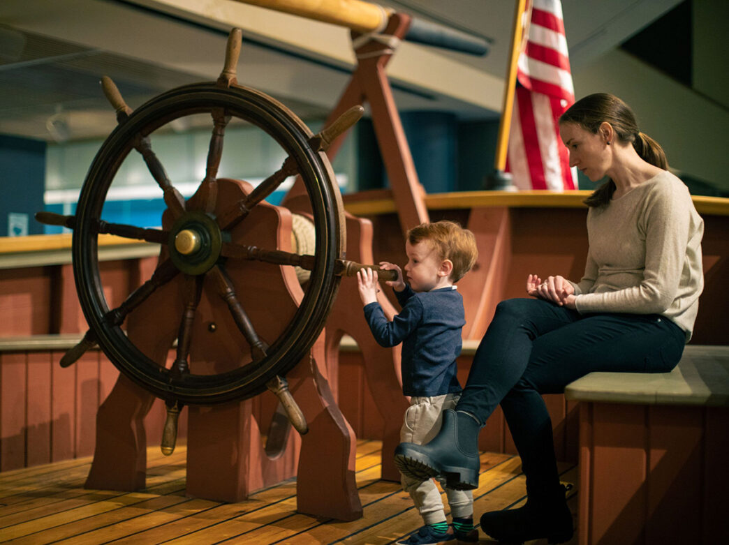 A toddle plays with a large ship wheel inside a nautical-themed exhibit while a woman sits behind them watching.