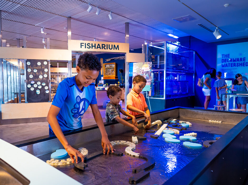 Three kids play with floating pieces in water table within the River Alive exhibit at the Independence Seaport Museum.