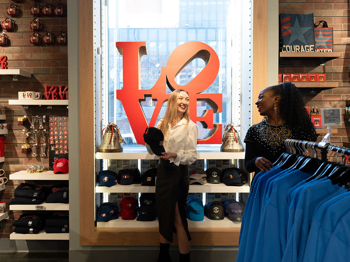 Two women browse Philadelphia-themed souvenirs in the brightly lit Independence Gift Shop, with a red LOVE sculpture in the background.