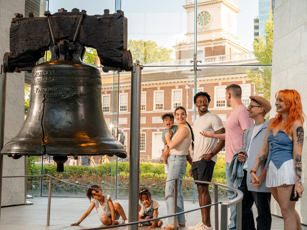 A group of visitors, including children and adults, admire the Liberty Bell inside Liberty Bell Center, with Independence Hall visible through the glass windows behind them.