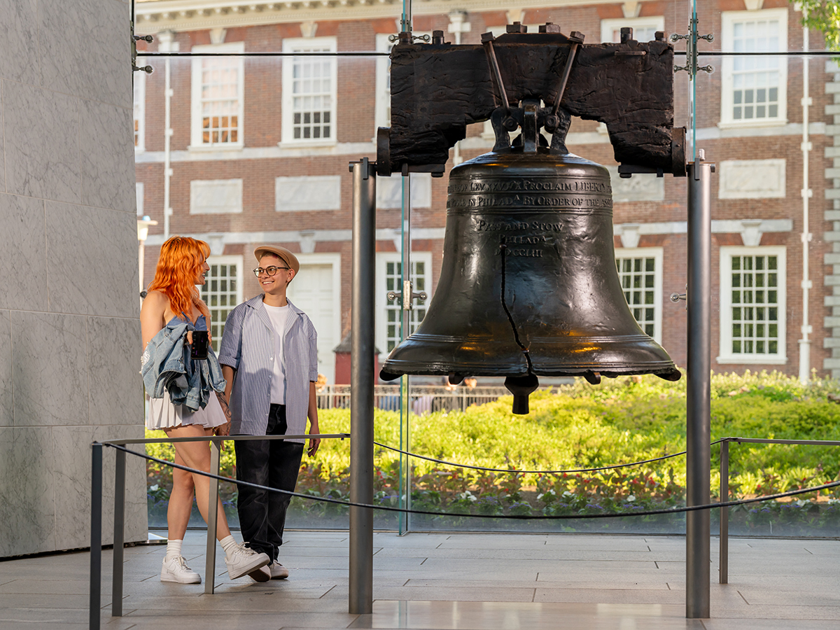 Two visitors smile and chat while standing next to the iconic Liberty Bell, with Independence Hall visible through the window behind them.