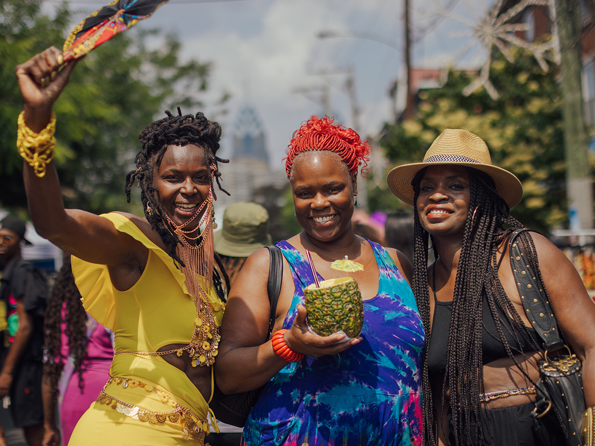 Three women smile for the camera at the ODUNDE Festival, one holding a pineapple drink and another waving a colorful fan.