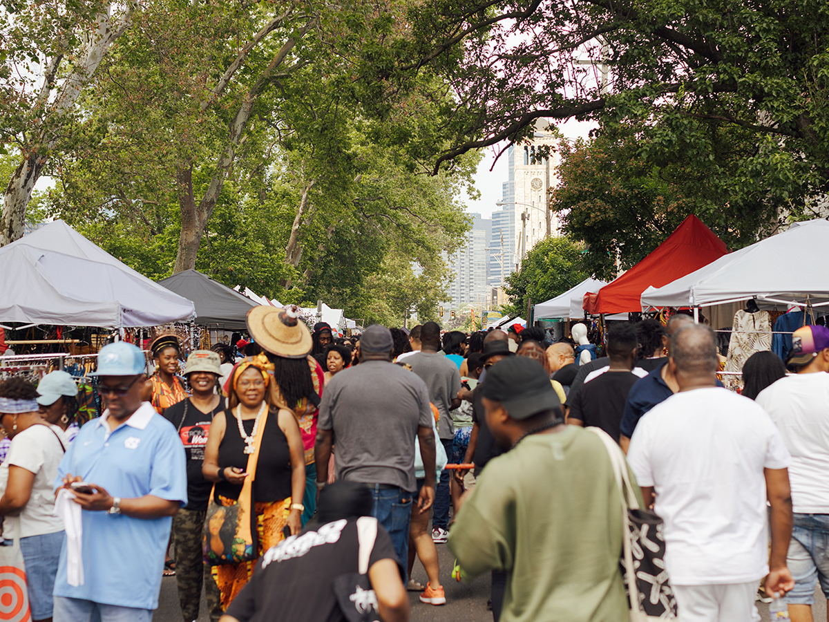 A bustling festival scene with people walking between vendor tents under shady trees with the Philadelphia skyline in the distance.