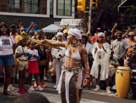 A dancers wearing a white bikini top and skirt performs int he middle of a crowd during the Odunde Festival, surrounded by spectators and live drummers.