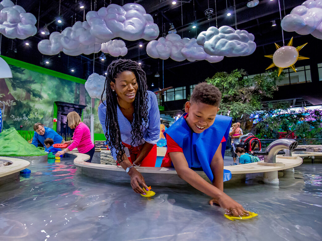 A smiling child and adult play with toy boats in an indoor water table at the Please Touch Museum. Hanging from the ceiling are fluffy clouds and a sun.