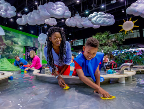 A smiling child and adult play with toy boats in an indoor water table at the Please Touch Museum. Hanging from the ceiling are fluffy clouds and a sun.