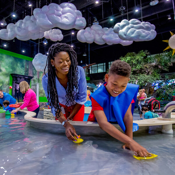 A smiling child and adult play with toy boats in an indoor water table at the Please Touch Museum. Hanging from the ceiling are fluffy clouds and a sun.