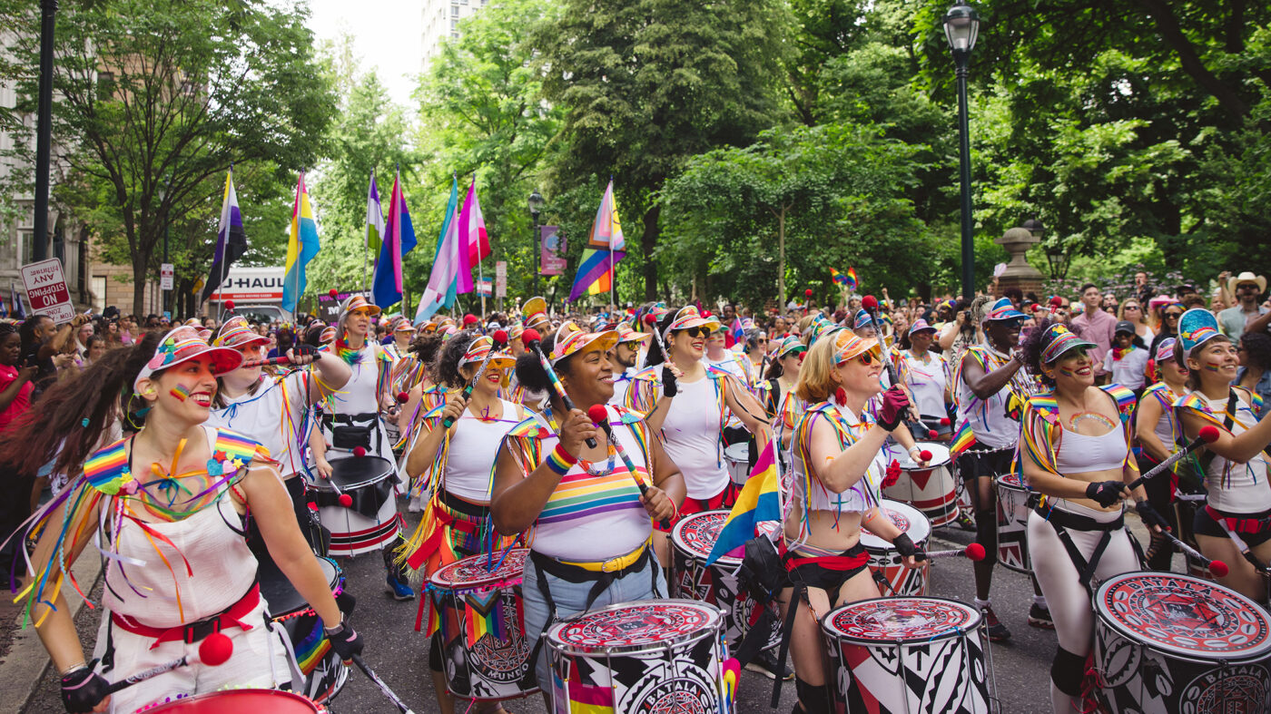 A joyful group of drummers in rainbow-themed outfits and visors parade down a city street, surrounded by waving Pride flags and cheering crowds.