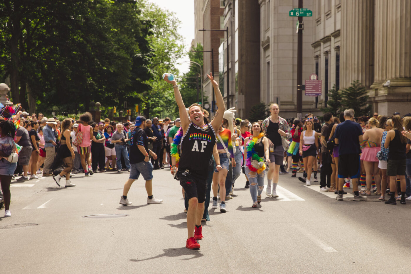 A festivalgoer dressed in a black outfit with the words "Proud AF" printed in rainbow-colored letters joyfully throws his hands in the air as he walks down a crowded street.