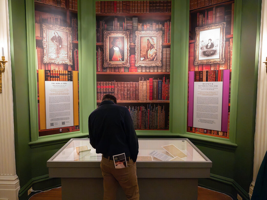 A museum visitor leans in to read historic texts in a glass display beneath portraits at The Rosenbach.