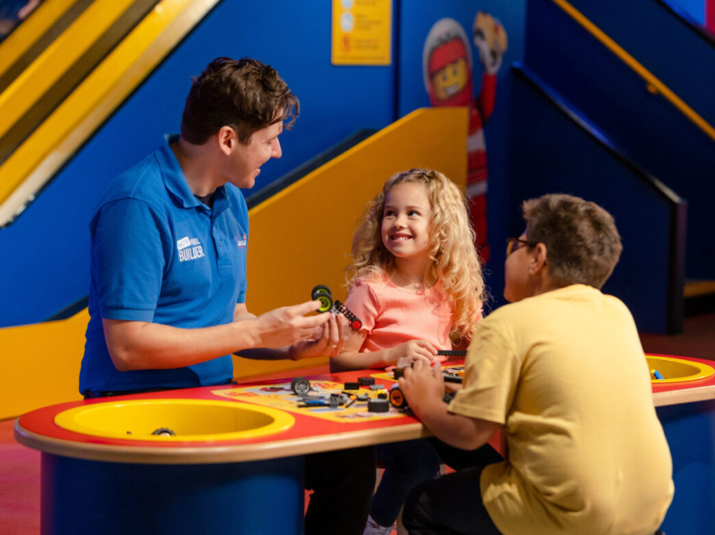 Two children sit at a building table while a staff member helps them build with Legos.