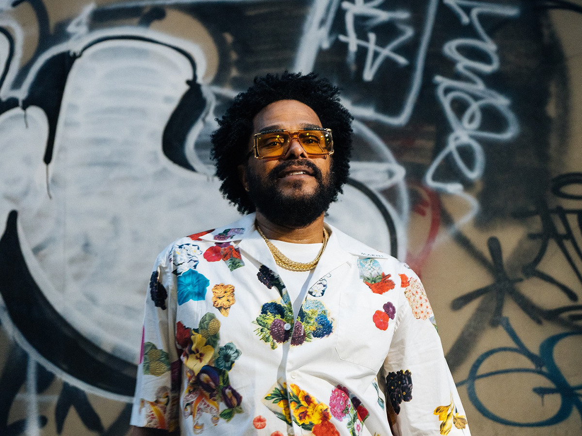 Singer-song writer, Maxwell, faces the camera while wearing amber sunglasses, a gold necklace and floral shirt. Behind him is wall covered in graffiti.