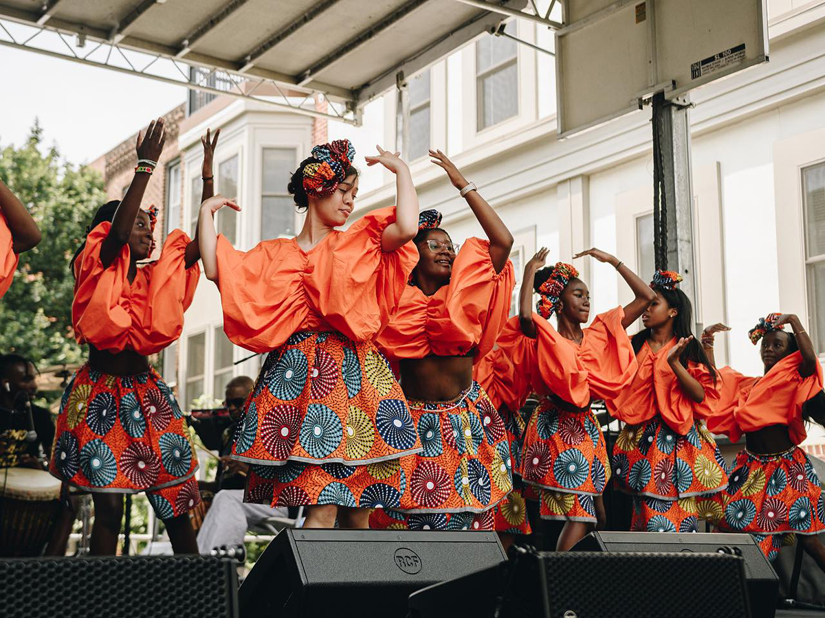 A group of dancers in matching outfits of orange poofy tops and vibrant patterned skirts perform on stage during the ODUNDE Festival.