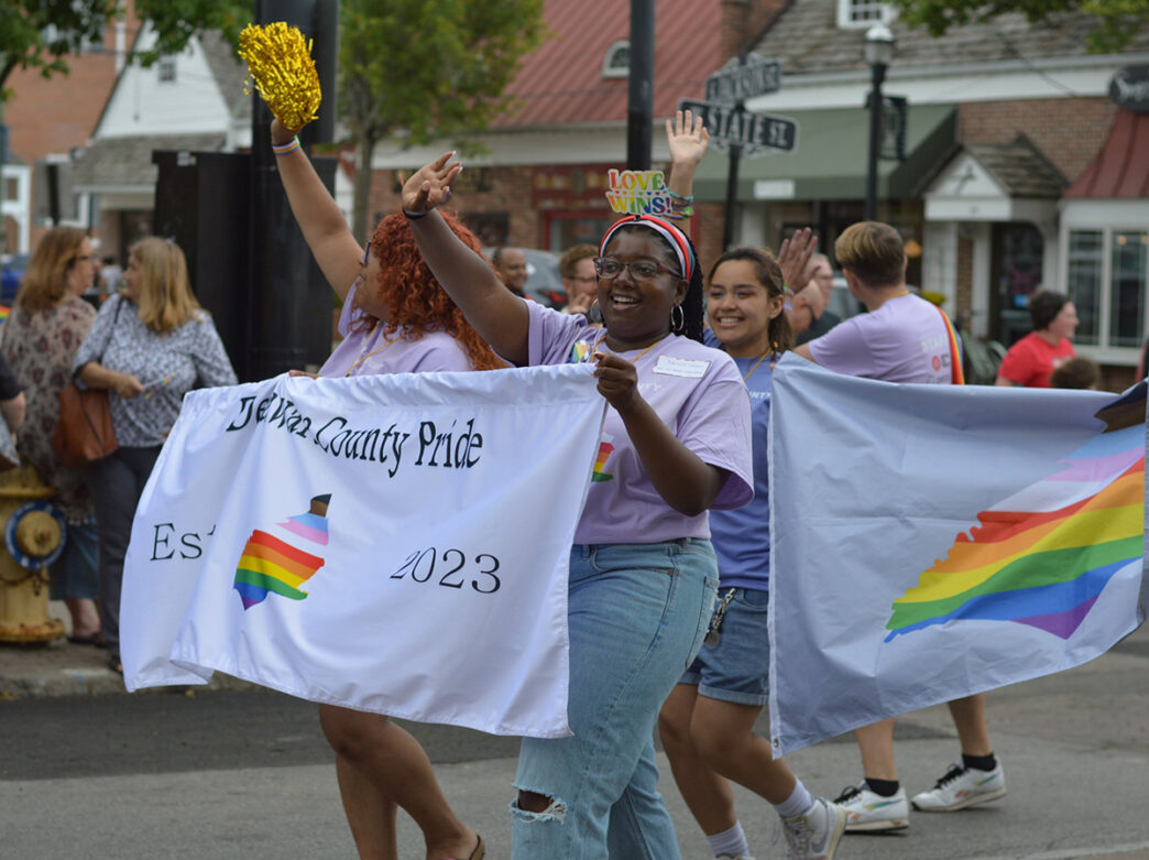 Des participants souriants, vêtus de chemises lavande, défilent lors de la Delaware County Pride Parade, agitant des pompons et tenant des banderoles décorées d'arc-en-ciel.