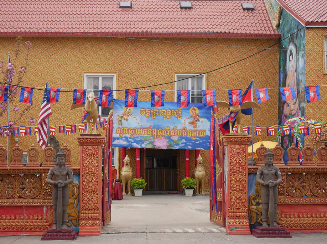 The exterior the Preah Buddha Rangsey Temple in Philadelphia, with a red and orange gate entrance decorated with bronze and gold statues.