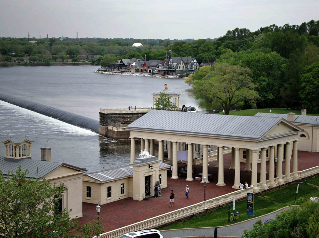 A neoclassical building with columns sits along the Schuylkill River, with a view of the dam and Boathouse Row in the background.
