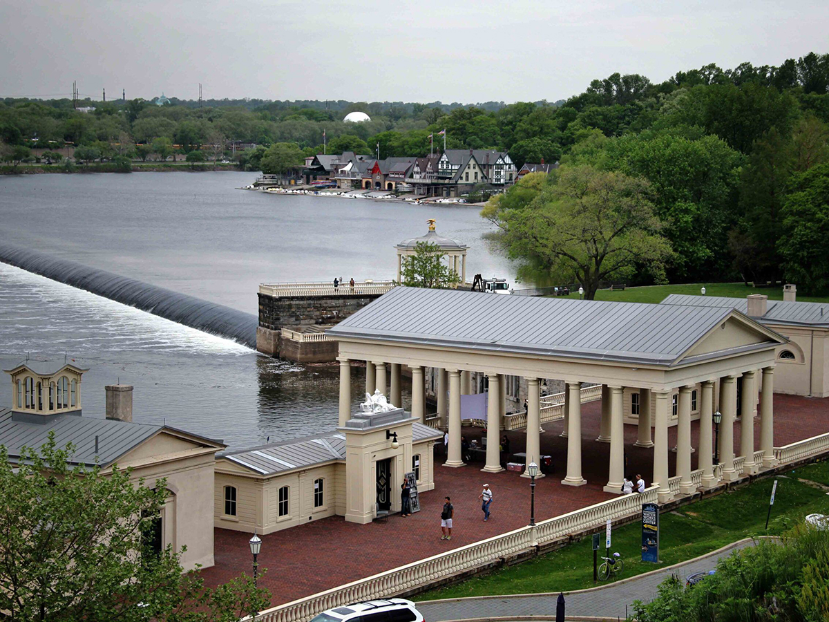 A neoclassical building with columns sits along the Schuylkill River, with a view of the dam and Boathouse Row in the background.