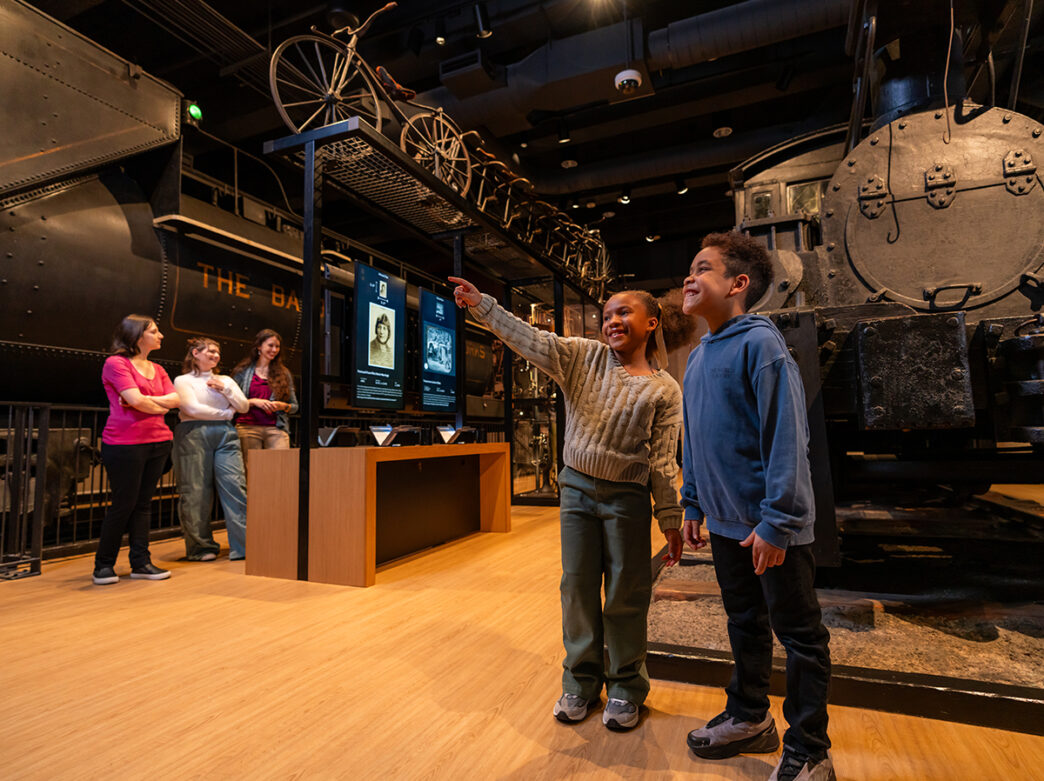 One child points toward something while another child looks in that direction and smiles. Behind them is a steam locomotive in the Hamilton Collections Gallery at The Franklin Institute.