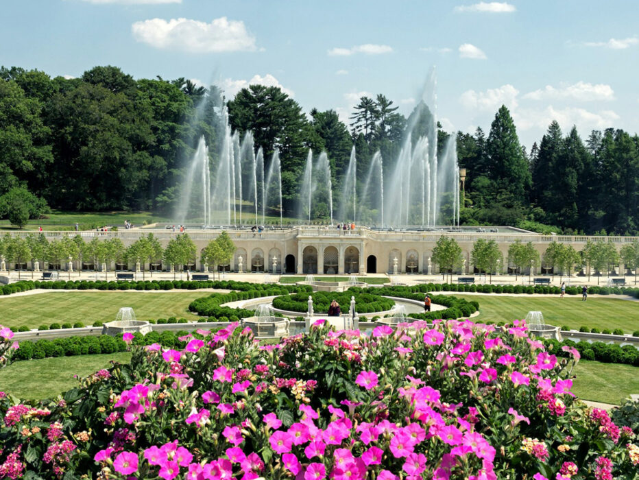 Fountains shoot water into the air above a grand formal garden at Longwood Gardens, framed by pink flowers in the foreground and lush trees in the background.