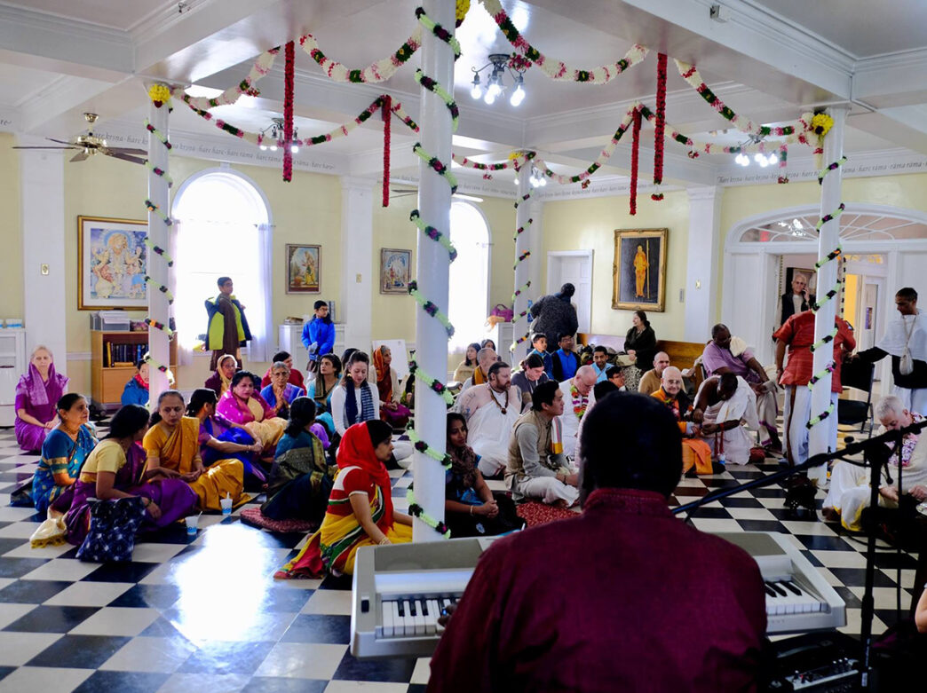 People sit cross-legged on the floor of a brightly lit temple, listening to live music played on a keyboard. The space is decorated with garlands of flowers.