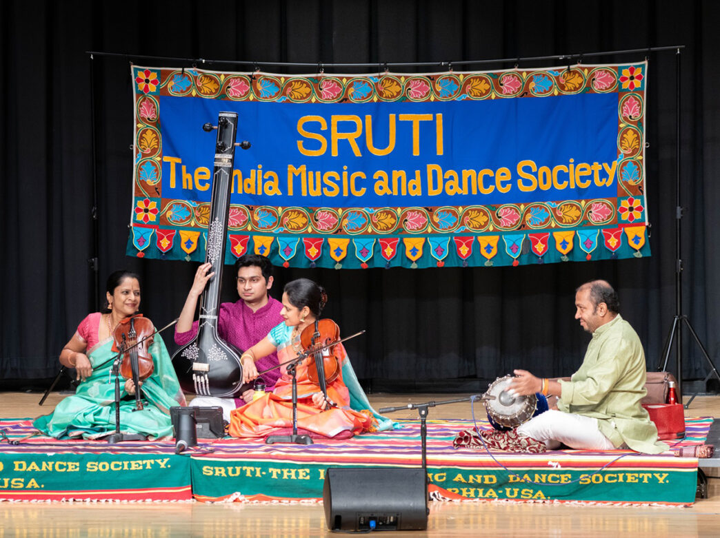 Four musicians sit on a rug while playing instruments, including a hand drum, violins and a sitar, in front of a colorful sign.