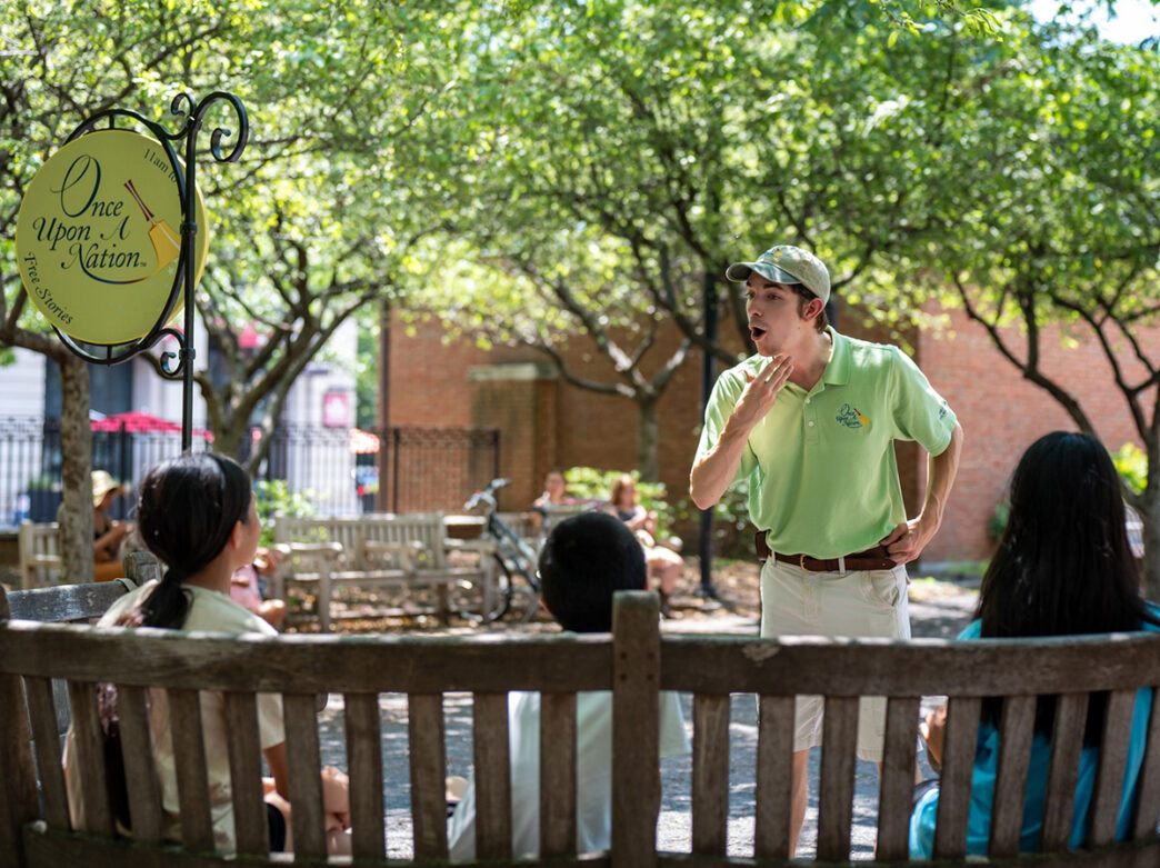 Three children sit on a bench outdoors while a storyteller performs in front of them.