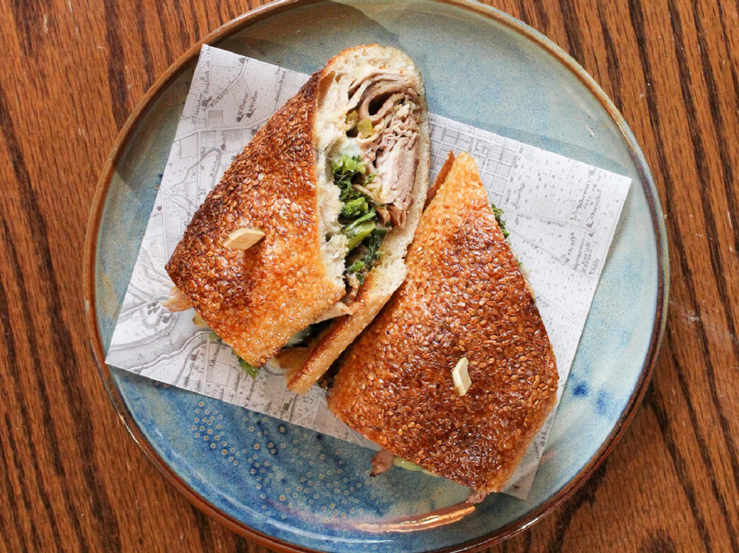 A roast pork sandwich cut in half is displayed on a blue plate on a wood table top. The roll is filled with layers of pork, greens and cheese.