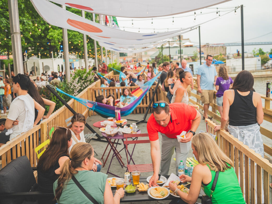 People eat at a table full of food and drinks, women sit on a colorful hammock and a crowd of people walk along the waterfront at Spruce Street Harbor Park