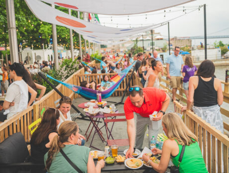 People eat at a table full of food and drinks, women sit on a colorful hammock and a crowd of people walk along the waterfront at Spruce Street Harbor Park