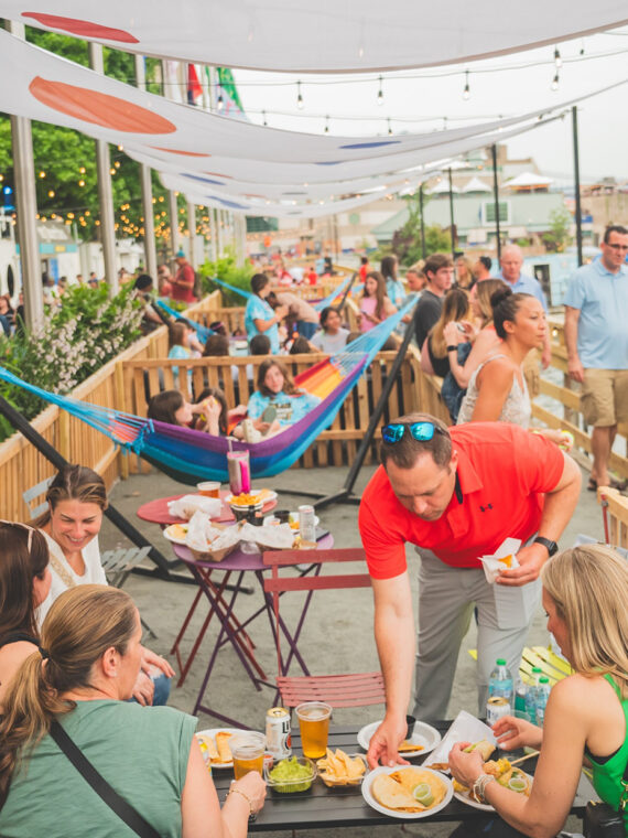 People eat at a table full of food and drinks, women sit on a colorful hammock and a crowd of people walk along the waterfront at Spruce Street Harbor Park