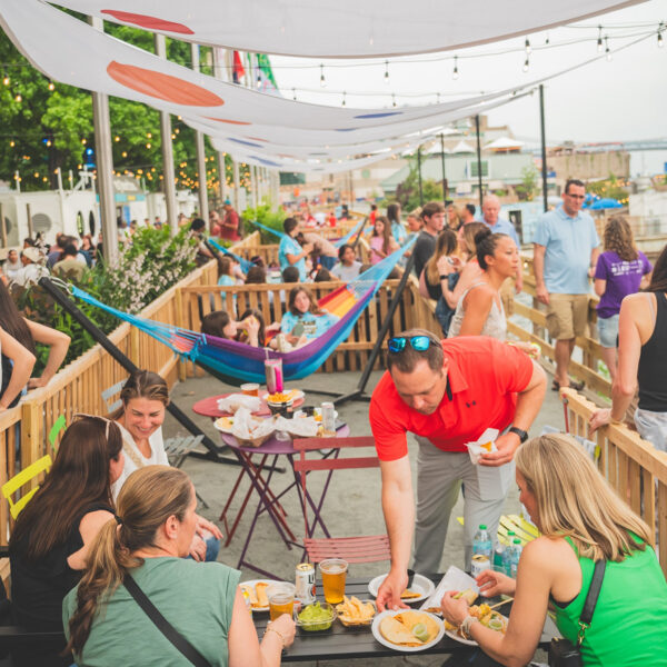 People eat at a table full of food and drinks, women sit on a colorful hammock and a crowd of people walk along the waterfront at Spruce Street Harbor Park