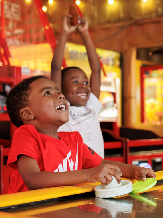 Surrounded by bright lights and colorful carnival games, two young boys laugh while playing air hockey at a Summerfest's lively arcade.