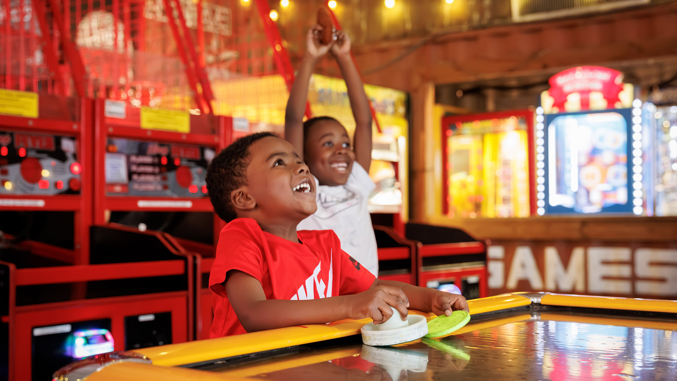 Surrounded by bright lights and colorful carnival games, two young boys laugh while playing air hockey at a Summerfest's lively arcade.