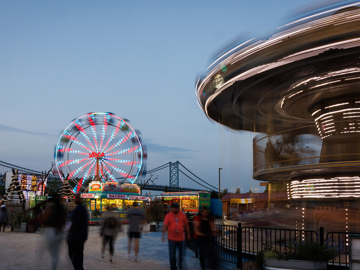 People walk through the Midway at Summerfest at dusk as the Ferris Wheel and Carousel are illuminated and in motion.
