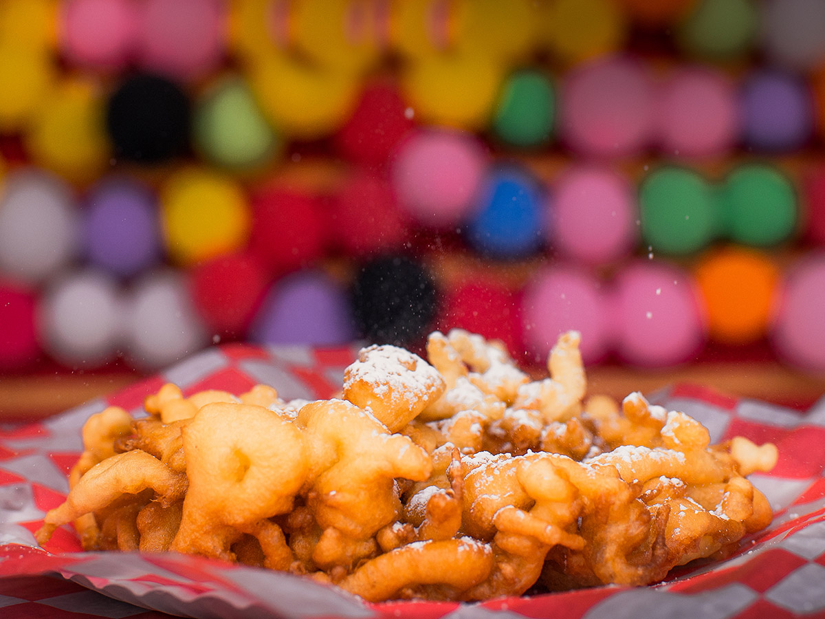 A delicious-looking funnel cake sits on red checkered paper as colorful carnival prizes can be seen in the background.