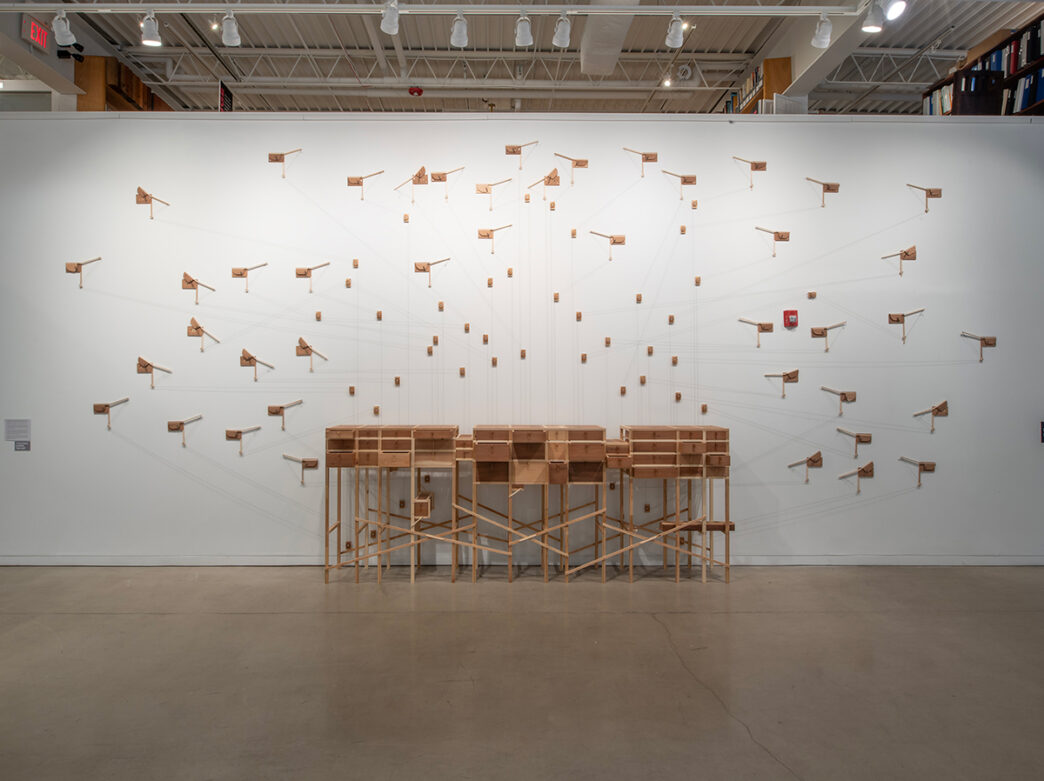A contemporary art installation featuring dozens of small wooden levers mounted on a white gallery wall, all connected to a wooden structure below with string.