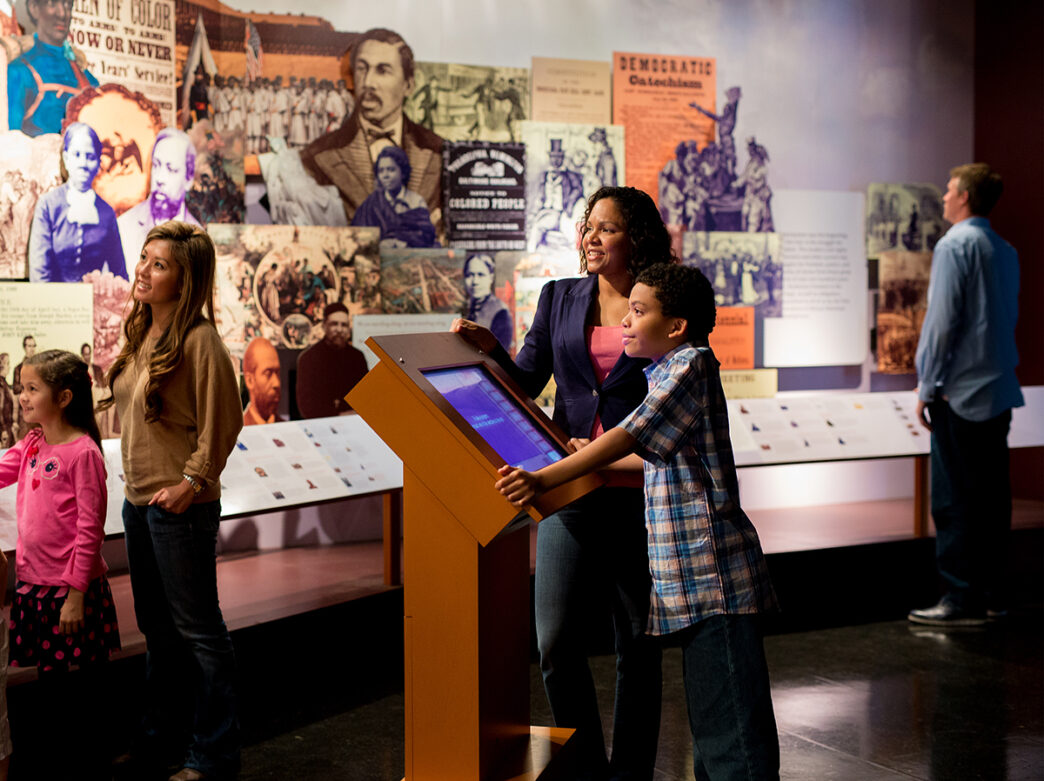 An adult and child stand behind a touch-screen display and look up at an exhibit at the African American Museum in Philadelphia.